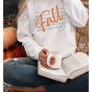 May include: A white sweatshirt with the word "Fall" written in orange script. The word is surrounded by a list of fall-related words in gray text, including "cozy", "sweater", "leaves", "apple cider", "bonfire", "autumn", "football", "starves", "apples", "boo", "harvest", "pumpkins", "pies", "caramel", "corn", and "coffee lattes".
