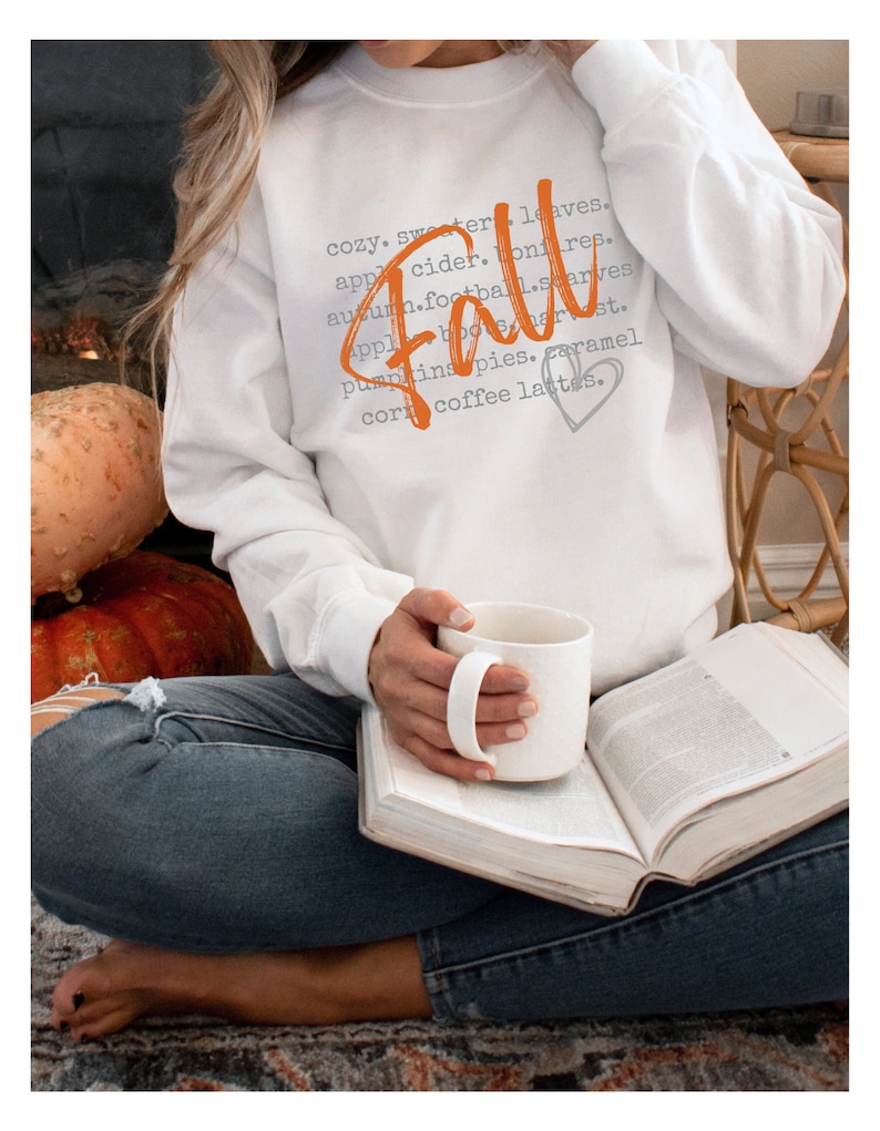 May include: A white sweatshirt with the word "Fall" written in orange script. The word is surrounded by a list of fall-related words in gray text, including "cozy", "sweater", "leaves", "apple cider", "bonfire", "autumn", "football", "starves", "apples", "boo", "harvest", "pumpkins", "pies", "caramel", "corn", and "coffee lattes".