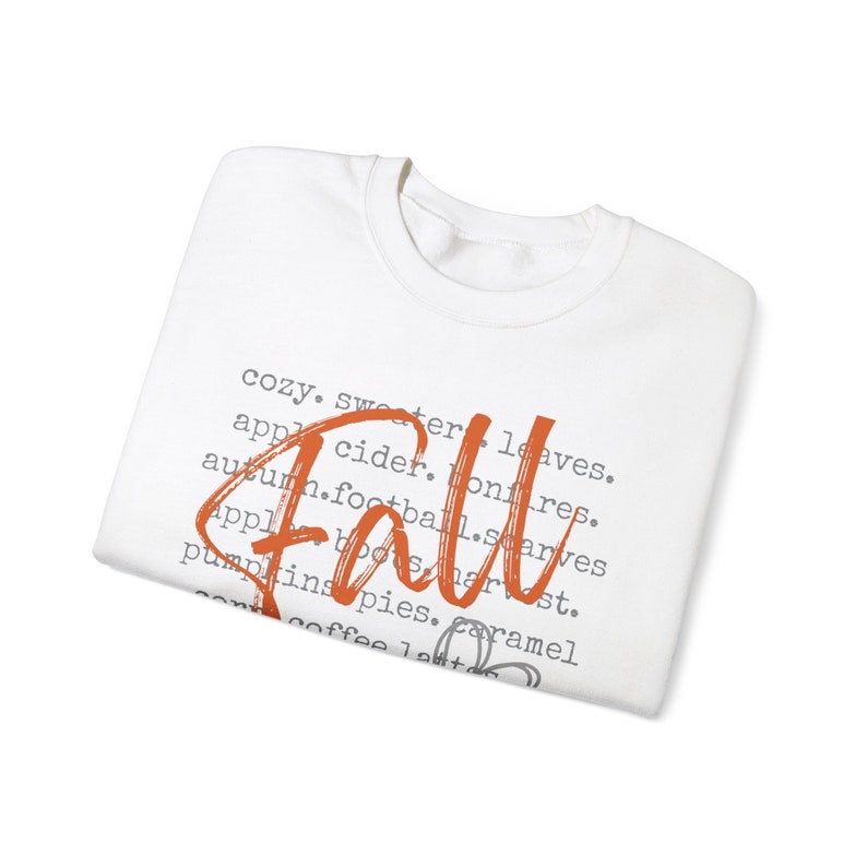 May include: White long-sleeve shirt with the word "Fall" written in orange script. The shirt has a list of words associated with the season, including "cozy", "sweater", "leaves", "apple cider", "bonfires", "autumn", "football", "scarves", "apple boots", "harvest", "pumpkins", "pies", "caramel", "coffee", and "latte".