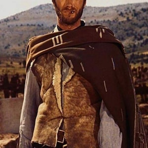May include: A man in a brown hat and poncho, with a brown vest over a light blue shirt. The image is a still from a Western film, with a mountainous backdrop.