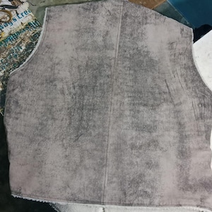 May include: A gray vest with a textured, suede-like exterior. The vest has a vertical seam down the center and a white, fuzzy lining visible at the bottom edge. The garment appears to be unlined.