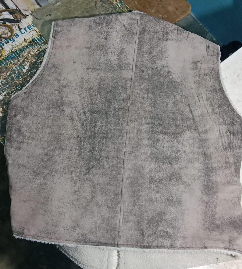 May include: A gray vest with a textured, suede-like exterior. The vest has a vertical seam down the center and a white, fuzzy lining visible at the bottom edge. The garment appears to be unlined.