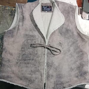 May include: A gray suede vest with a white, fluffy interior lining and collar. The vest has a tie closure and a label that reads "Real Leather". The vest is a neutral color and appears to be made of a soft material.