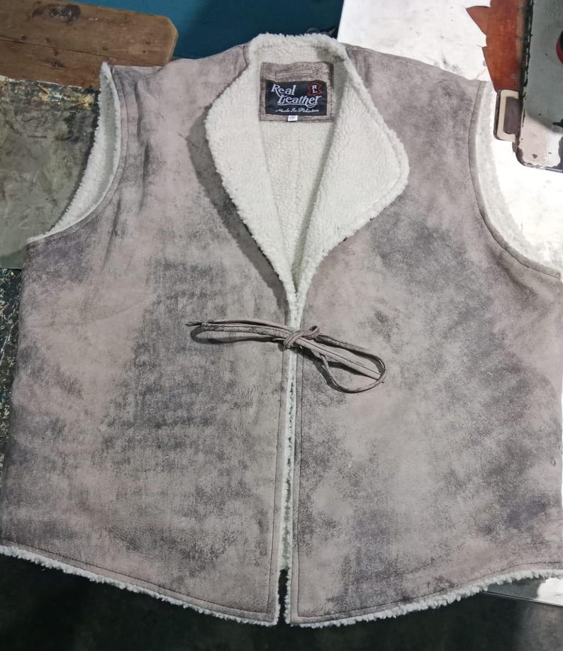May include: A gray suede vest with a white, fluffy interior lining and collar. The vest has a tie closure and a label that reads "Real Leather". The vest is a neutral color and appears to be made of a soft material.