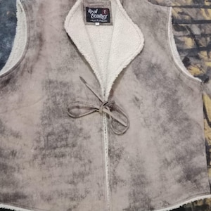 May include: A tan leather vest with a cream-colored, fuzzy lining and collar. The vest has a tie closure and a label that reads "Real Leather". The vest has a distressed, mottled appearance.
