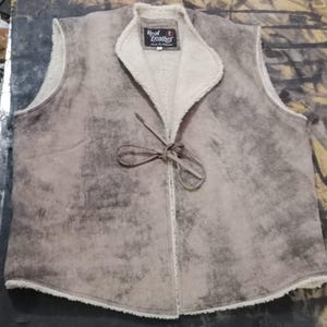 May include: A tan and gray suede vest with a cream-colored, fuzzy lining and collar. The vest has a tie closure and a label that reads "Real Leather". The vest is sleeveless and appears to be made of a soft material.