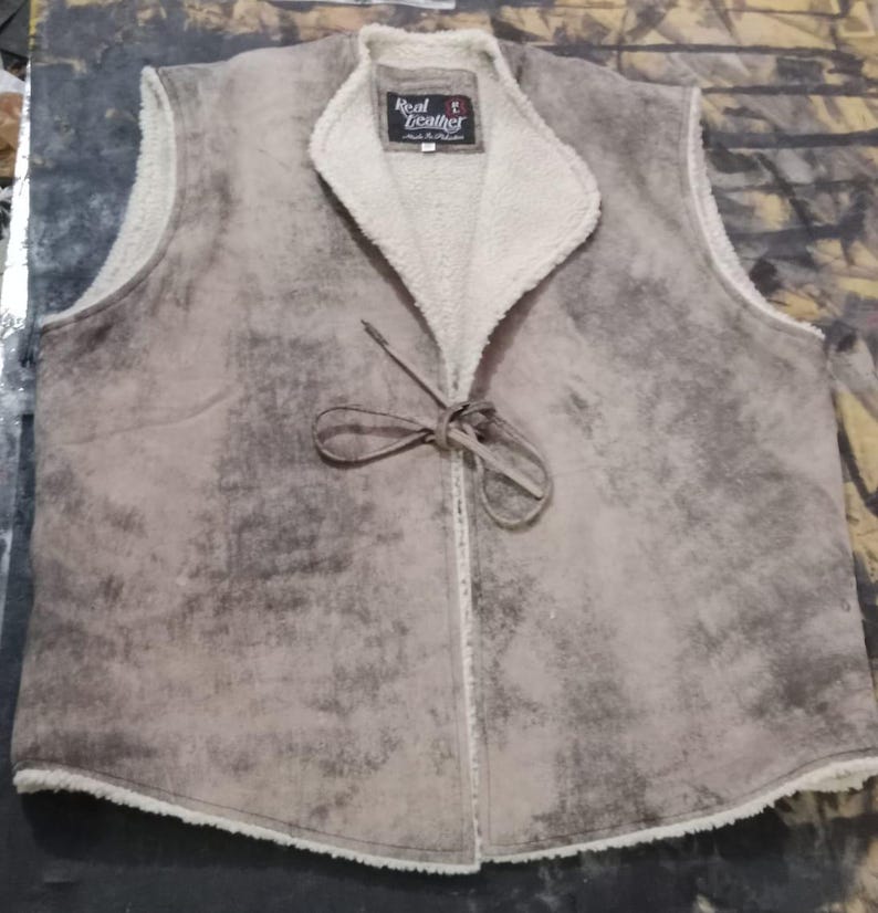 May include: A tan and gray suede vest with a cream-colored, fuzzy lining and collar. The vest has a tie closure and a label that reads "Real Leather". The vest is sleeveless and appears to be made of a soft material.