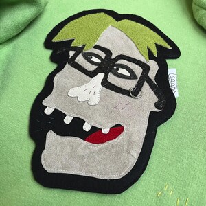 May include: A lime green hoodie featuring a felt applique of a cartoon face with glasses, green hair, and a red mouth. The applique is outlined in black and has a small tag on the side. The hoodie has a kangaroo pocket.
