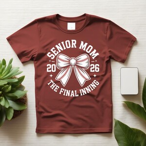 Może przedstawiać: Bordowa koszulka z białym motywem baseballowym. Grafika zawiera napisy "Senior Mom 2026" i "The Final Inning" otaczające kokardę w kształcie piłki do baseballu. Koszulka jest prezentowana na białej powierzchni.