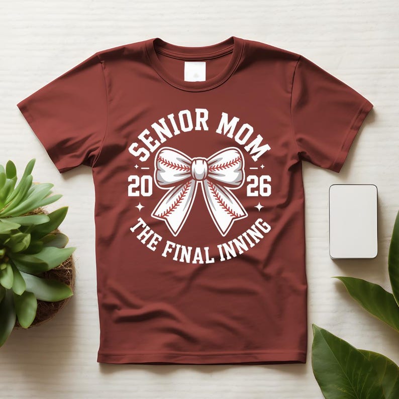 Może przedstawiać: Bordowa koszulka z białym motywem baseballowym. Grafika zawiera napisy "Senior Mom 2026" i "The Final Inning" otaczające kokardę w kształcie piłki do baseballu. Koszulka jest prezentowana na białej powierzchni.