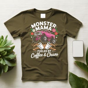 May include: Olive green t-shirt featuring a pink monster truck graphic with the words "Monster Mama" and "Fueled by Coffee & Chaos." The design includes flowers and coffee cups, creating a fun and energetic vibe.