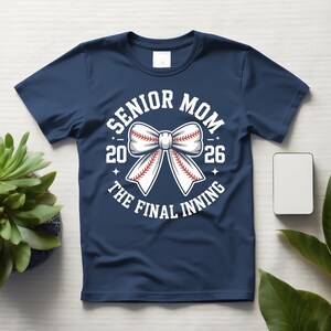 Może przedstawiać: Granatowa koszulka z białym motywem baseballowym. Projekt zawiera napis "SENIOR MOM 2026 THE FINAL INNING" otaczający kokardę w kształcie piłki do baseballu.