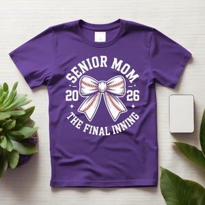 Może przedstawiać: Fioletowa koszulka z białym nadrukiem z napisem "Senior Mom 2026 The Final Inning". Grafika przedstawia kokardę w stylu baseballowym. Koszulka jest prezentowana na jasnej powierzchni z rośliną i małym białym prostokątem.