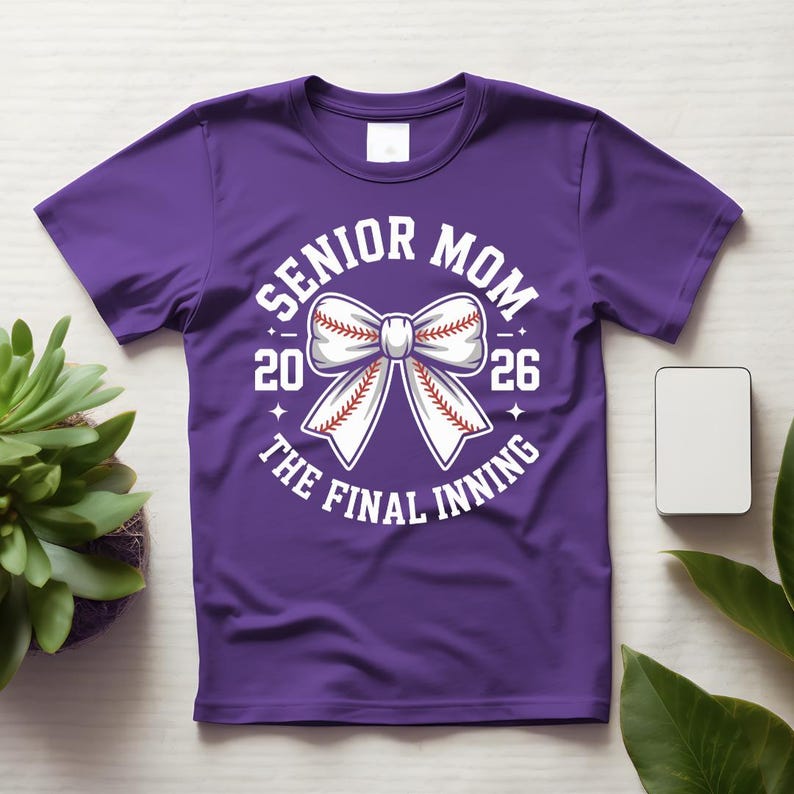 Może przedstawiać: Fioletowa koszulka z białym nadrukiem z napisem "Senior Mom 2026 The Final Inning". Grafika przedstawia kokardę w stylu baseballowym. Koszulka jest prezentowana na jasnej powierzchni z rośliną i małym białym prostokątem.