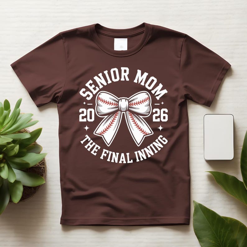 Może przedstawiać: Bordowa koszulka z białą grafiką przedstawiającą kokardę w stylu baseballowym oraz napisem "SENIOR MOM 2026 THE FINAL INNING". Projekt zawiera piłkę do baseballu z czerwonymi szwami.