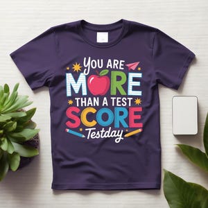 May include: A dark purple t-shirt with the text "You are More Than a Test Score Testday" in colorful letters and a red apple graphic. The design includes stars, pencils, and a paper airplane.