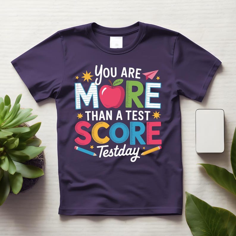 May include: A dark purple t-shirt with the text "You are More Than a Test Score Testday" in colorful letters and a red apple graphic. The design includes stars, pencils, and a paper airplane.