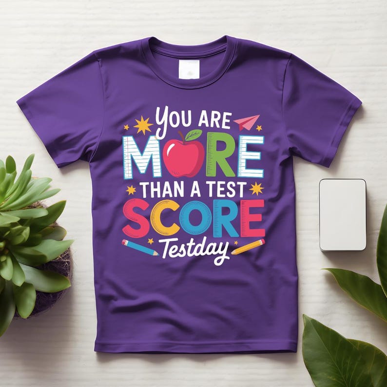 May include: A purple t-shirt with the text "You are More Than a Test Score Testday" in colorful letters. The design includes an apple, pencils, and stars. The shirt is made of a soft material.