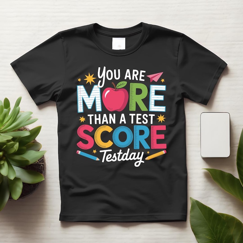 May include: Black t-shirt with the text "You Are More Than A Test Score Testday" in colorful letters and a red apple graphic. The shirt has a crew neck and short sleeves. The design includes pencils and paper airplanes.