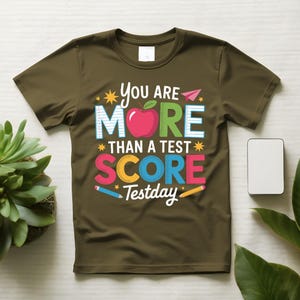 May include: Olive green t-shirt with the text "You Are More Than A Test Score Testday". The design includes a red apple, colorful text, and pencil illustrations. The shirt is a crew neck.