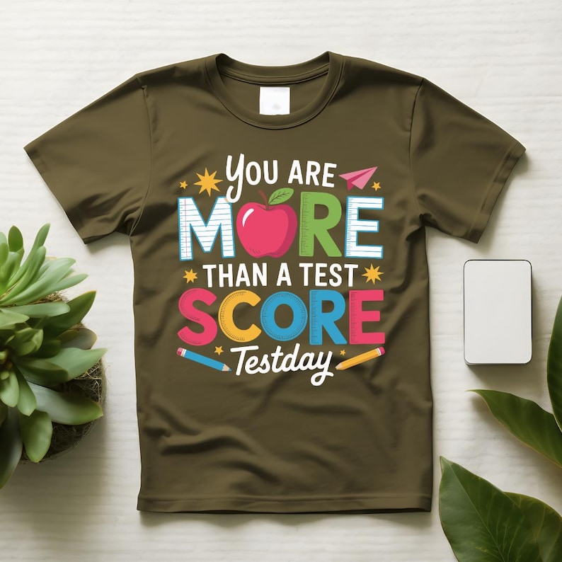 May include: Olive green t-shirt with the text "You Are More Than A Test Score Testday". The design includes a red apple, colorful text, and pencil illustrations. The shirt is a crew neck.