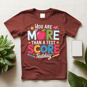 May include: A maroon t-shirt with the text "You Are More Than A Test Score Testday" in colorful letters and graphics. The design includes an apple, pencils, and stars, suitable for students and teachers.