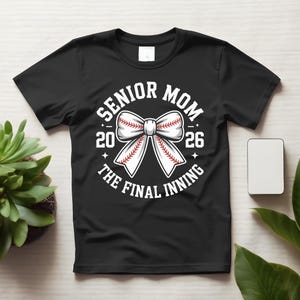 Może przedstawiać: Czarna koszulka z białym wzorem kokardy w stylu baseballowym. Tekst brzmi "SENIOR MOM 2026 THE FINAL INNING". Projekt zawiera piłkę do baseballu z czerwonymi szwami i gwiazdami.