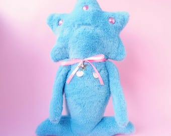 Può includere: Un peluche a forma di stella blu chiaro con un nastro rosa e un ciondolo argentato. La stella ha accenti di gemme rosa. Il giocattolo è su uno sfondo rosa.