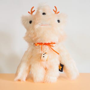 Op de afbeelding: Een crèmekleurig pluche wezen met lange, ruige vacht, drie zwarte ogen en twee kleine oranje geweien. Het draagt een oranje lint om de nek met een kleine, beige, kopvormige bedel die aan een wit koord hangt en een zwart label aan de zijkant.
