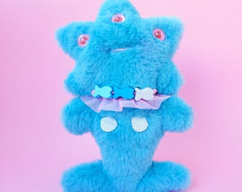 Può includere: Un peluche turchese a forma di stella, con tre occhi rosa a forma di bottone. Ha un colletto a balze ornato con decorazioni a forma di pesce e due accenti bianchi smerlati. Il giocattolo è ambientato su uno sfondo rosa.
