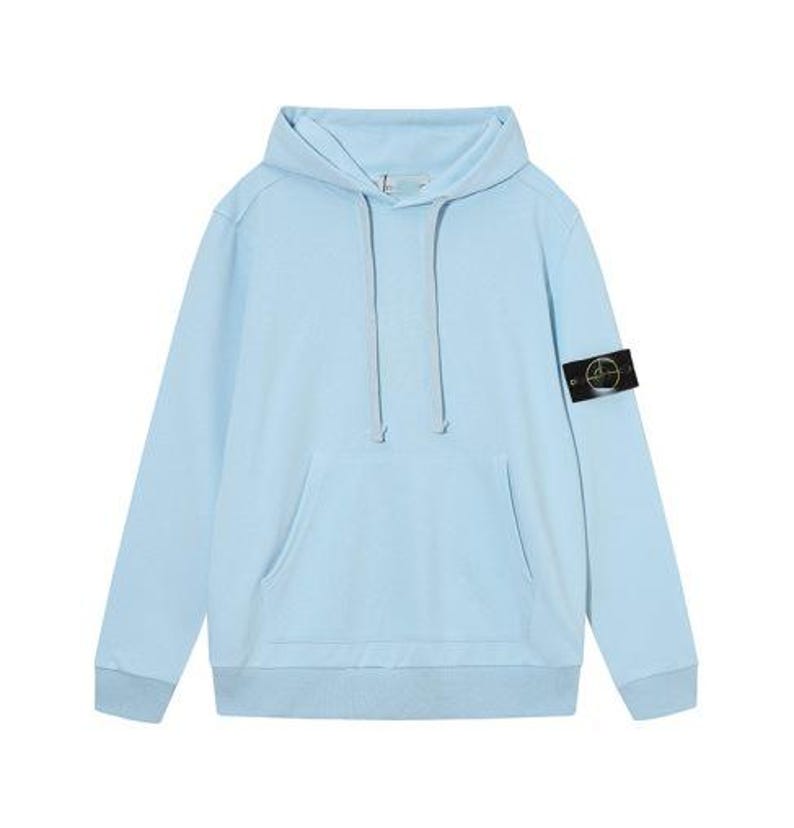 May include: A light blue hoodie with a front pocket and a drawstring hood. The long-sleeved sweatshirt features a small black and gold patch on the left sleeve. The garment is made of a soft material.