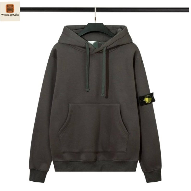 May include: A dark gray hoodie with a drawstring hood and a kangaroo pocket. The sweatshirt has a patch on the left sleeve. The hoodie is hanging on a black hanger.