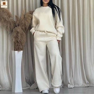 Puede incluir: Conjunto de ropa de estar por casa color crema, con una sudadera de cuello redondo y pantalones de pernera ancha. La sudadera tiene mangas largas y los pantalones tienen cintura con cordón. El conjunto está hecho de un material suave y cómodo.