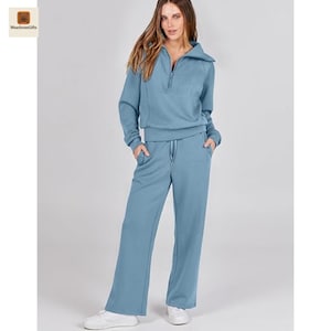 May include: A two-piece casual wear set in a muted blue hue. The top is a long-sleeved sweatshirt with a zip and a high collar. The trousers are wide-legged with a drawstring. The model is wearing white trainers.