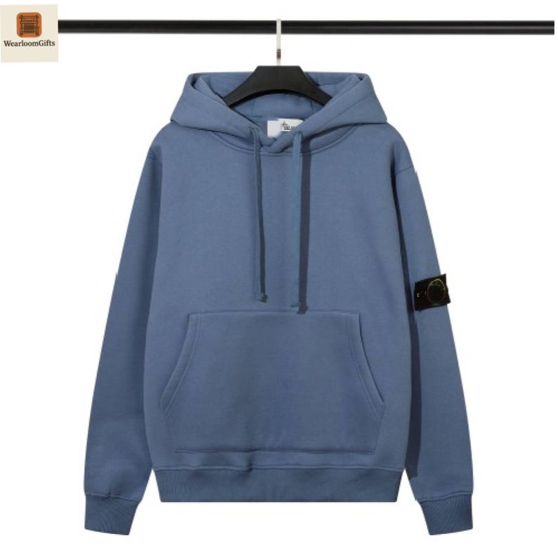 May include: A solid blue hoodie with a kangaroo pocket and a drawstring hood. The sweatshirt has a small patch on the left sleeve. The hoodie is hanging on a black hanger.