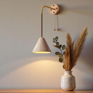 Könnte beinhalten: Eine Wandleuchte mit einem konischen Schirm. Die Leuchte hat einen Holzsockel, einen Messingarm und eine Zugkette. Der Schirm ist hellbeige und die Innenseite goldfarben. Die Leuchte ist an einer hellgrauen Wand befestigt.
