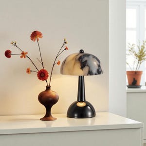 May include: A contemporary table lamp with a black base and stem, featuring a marbled, off-white and grey dome-shaped shade. A small, round, black knob is on top. A wooden vase with orange flowers is to the left.