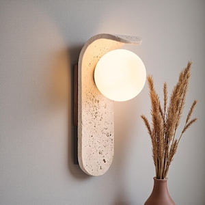 May include: A modern wall sconce with a textured, off-white finish and a spherical, glowing light. The sconce has a curved design and is mounted on a dark wooden backing. A vase of dried plants sits nearby.
