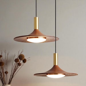 May include: Two pendant lights featuring wide, circular wooden shades with a dark brown grain. Each fixture has a cylindrical brass-toned top connector, a black suspension cord, and a central circular white light diffuser with a copper rim.