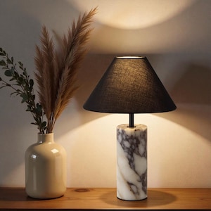 May include: A modern table lamp with a black lampshade and a marble base. The base is cylindrical with white and gray veining. The lamp sits on a wooden surface, next to a vase with dried plants.