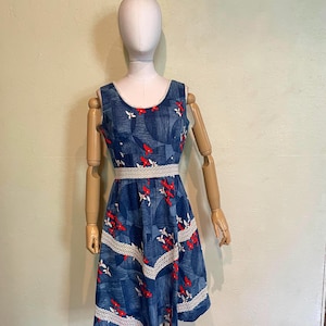 May include: A blue sleeveless dress with white lace trim and red floral details. The dress is displayed on a mannequin. It features a scoop neckline and a fitted waist. The skirt flares from the waist.