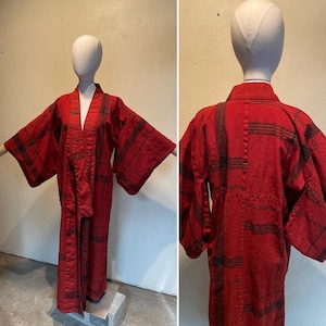 May include: A red kimono with black stripes is displayed on a mannequin. The kimono has wide sleeves and a wrap-around design. The fabric appears textured. The garment is a traditional Japanese robe.