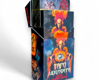 May include: A deck of tarot cards in a black box with a fiery, illustrated figure featuring a prominent eye on the front. The box displays the text "ТАРО ЛЮЦИФЕРА" in a stylized font, with additional Cyrillic text below and on the side flap.