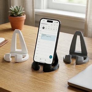 May include: A smartphone displaying a calendar app is propped up in a black phone stand. Two additional phone stands, one white and one gray, are also on the wooden surface. A notebook, pen, and a small potted plant are also visible.