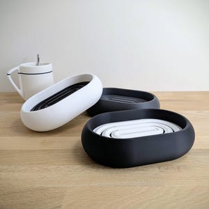 Può includere: Tre portasapone ovali bianchi, neri e grigi, con interno a coste. Sullo sfondo una tazza bianca con una striscia nera e un cucchiaino. I piatti sono su una superficie di legno chiaro.