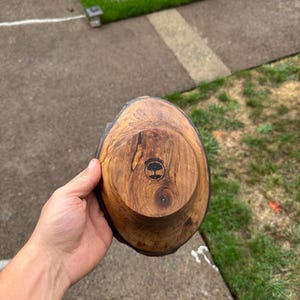 Pu&ograve; includere: Una ciotola di legno fatta a mano con un bordo naturale. La ciotola &egrave; realizzata in legno marrone scuro con un disegno circolare sul fondo. La ciotola &egrave; tenuta nella mano di una persona.