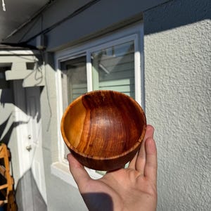 Pu&ograve; includere: Una ciotola di legno fatta a mano, di un ricco colore marrone caldo. La ciotola ha una finitura liscia e lucida e un motivo naturale di venature del legno. La ciotola &egrave; tenuta in una mano, mostrando la sua forma rotonda e le variazioni di tonalit&agrave; del legno.