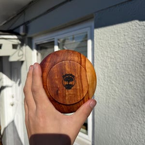 Pu&ograve; includere: Un contenitore rotondo in legno con coperchio. Il contenitore &egrave; realizzato in legno marrone chiaro con un motivo a venature pi&ugrave; scure. Il coperchio presenta un disegno di albero nero. Il contenitore &egrave; tenuto in una mano.