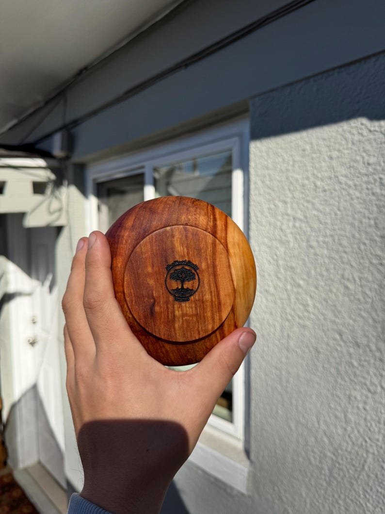 Pu&ograve; includere: Un contenitore rotondo in legno con coperchio. Il contenitore &egrave; realizzato in legno marrone chiaro con un motivo a venature pi&ugrave; scure. Il coperchio presenta un disegno di albero nero. Il contenitore &egrave; tenuto in una mano.