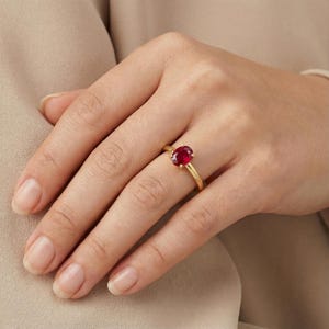 May include: A gold ring featuring a vibrant red oval gemstone. The ring's band is a simple, polished gold, and the gemstone is the focal point. The ring is displayed on a hand, showcasing its design and color.